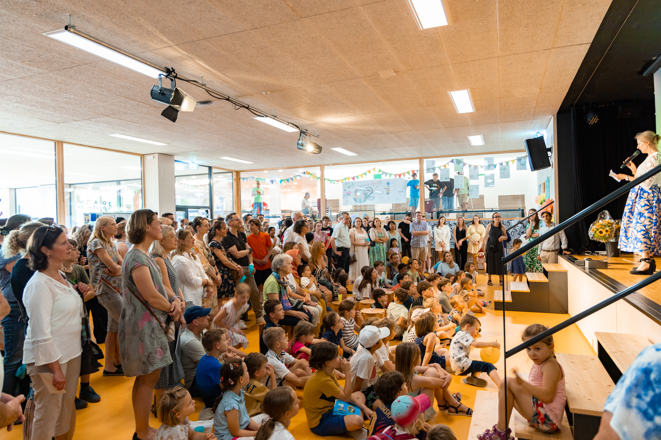 Kinder und Eltern in der Aula stehen und sitzen am Boden